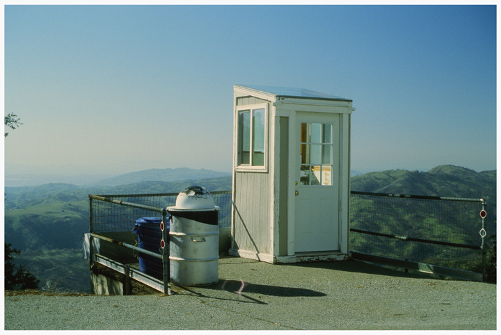 A small white booth and a trash can sit on a paved overlook with a vast mountainous landscape in the background.