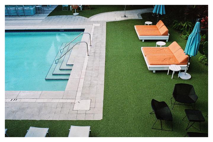 A swimming pool with steps and handrails is next to a green lawn with lounge chairs, umbrellas, and other seating.