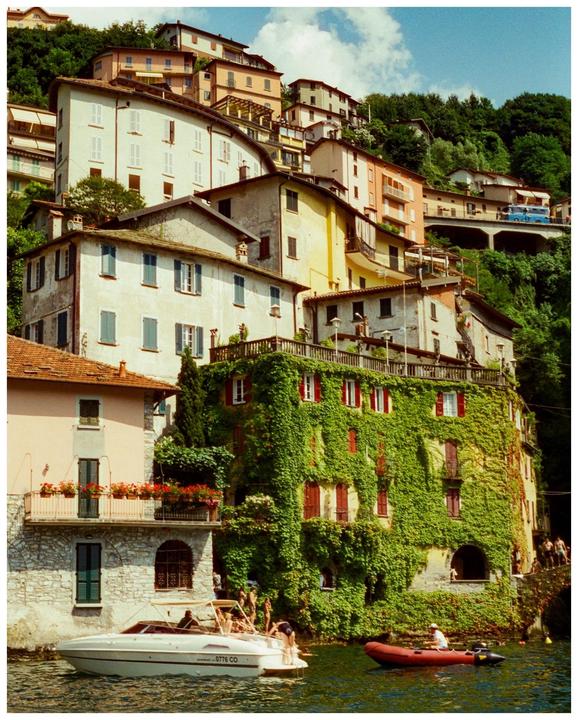 A hillside village with houses built into the slope, some covered in ivy, overlooks a body of water with boats.