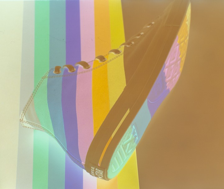 A shoe with rainbow stripes is angled against a background of vertical rainbow stripes.