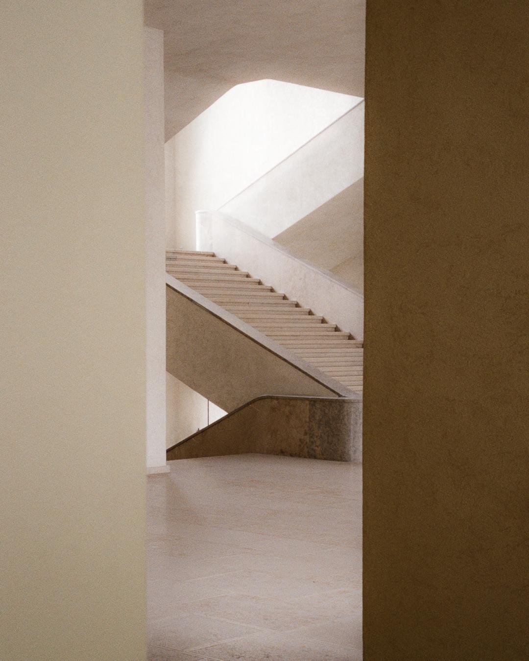 A minimalist architectural interior features a light-colored staircase with a curved railing, illuminated by natural light from above.