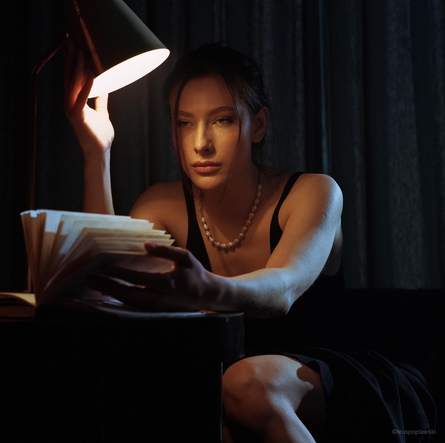 A woman wearing a pearl necklace sits at a table, looking at a stack of papers illuminated by a desk lamp.