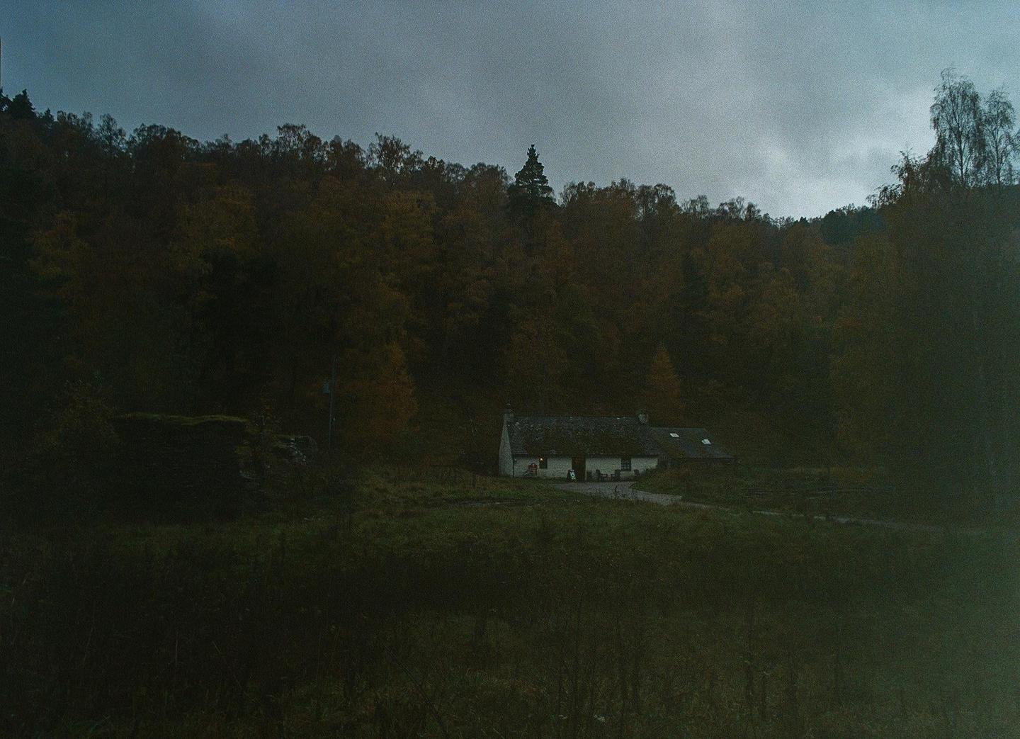 A small white house with a dark roof sits in a grassy field at the base of a forest under a cloudy sky.