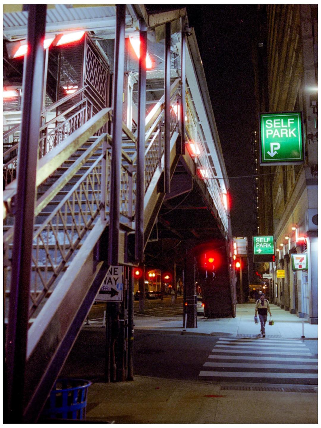 A person walks across a crosswalk at night next to a large metal structure and a "self park" sign.