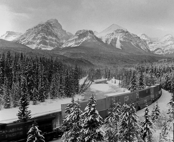 A long freight train moves through a snow-covered forest with towering mountains in the background.