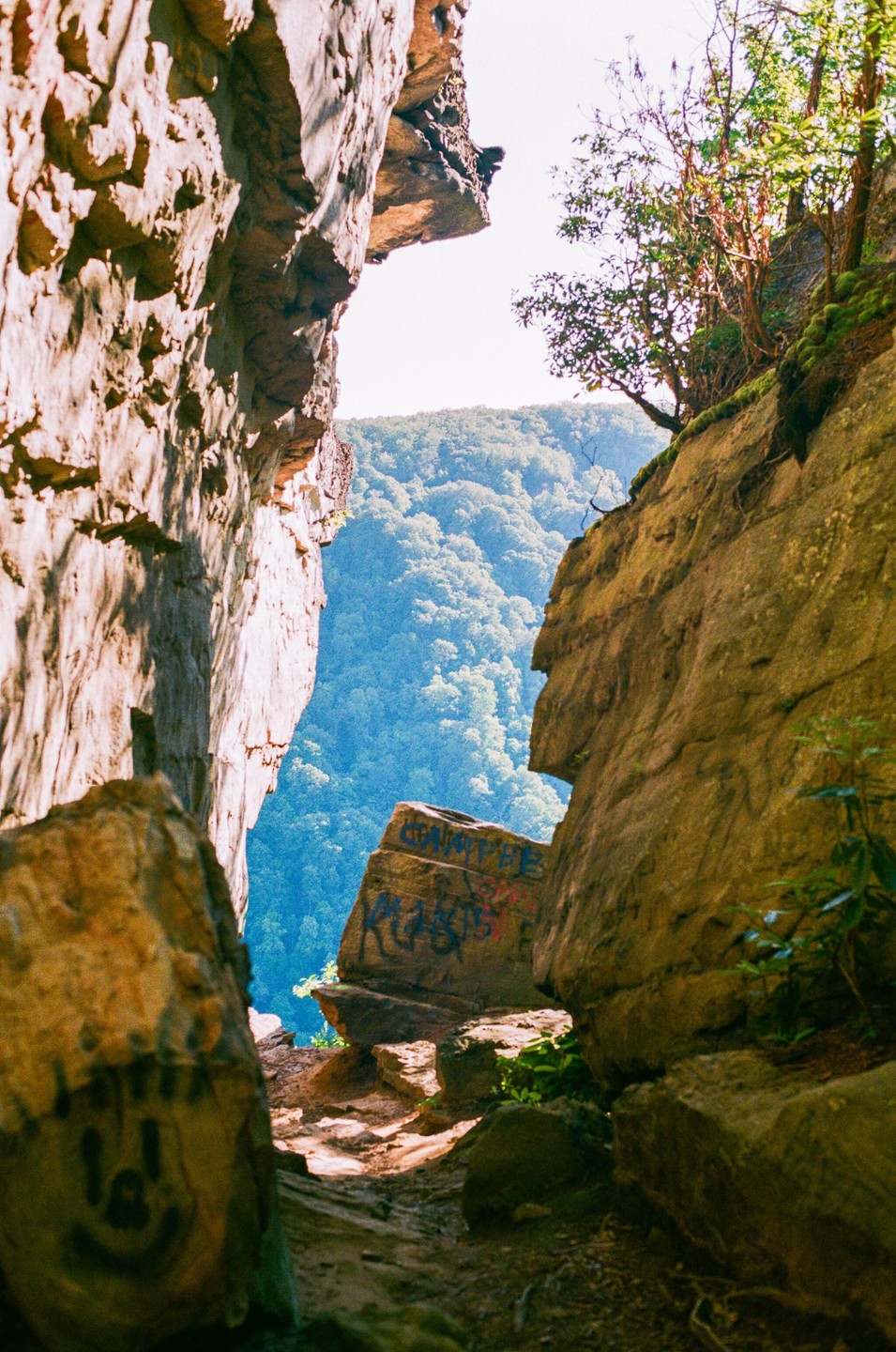 A narrow gap between two large rock formations reveals a lush, green valley in the distance under a bright sky.