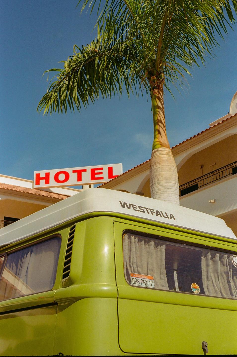A green Westfalia van is parked in front of a hotel sign and a palm tree.