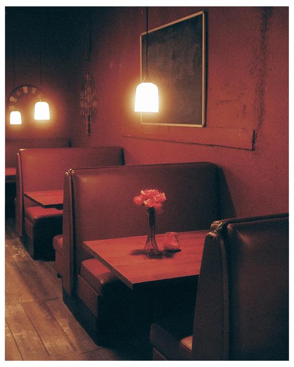 A dimly lit restaurant interior features red booths and tables, with a vase of pink flowers on one table.