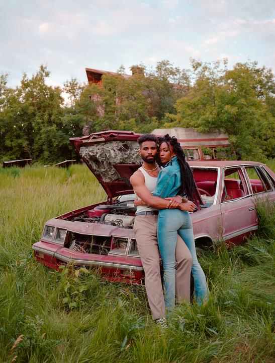 A couple embraces in front of a broken-down car in a grassy field.