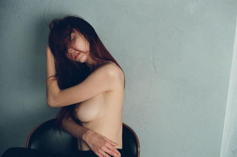 A woman with long red hair sits topless in a chair against a light gray wall.