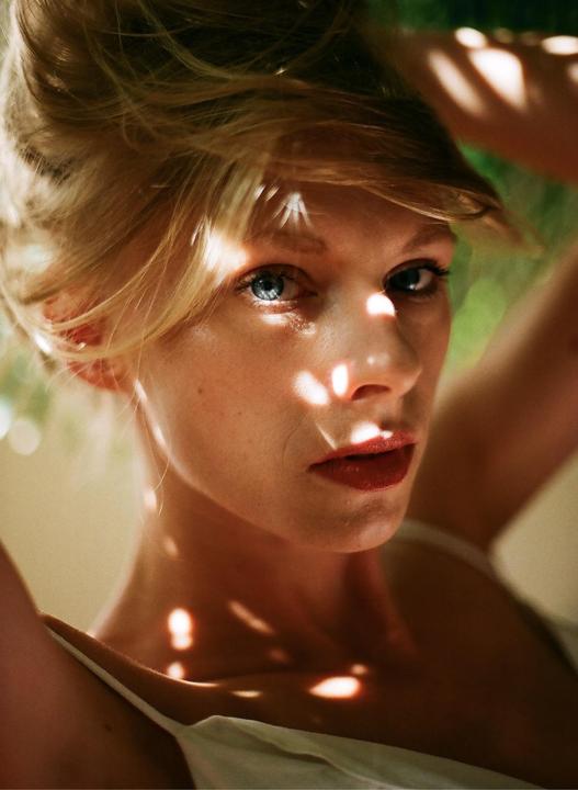 A woman with blonde hair and blue eyes looks directly at the viewer, with dappled sunlight on her face and shoulders.