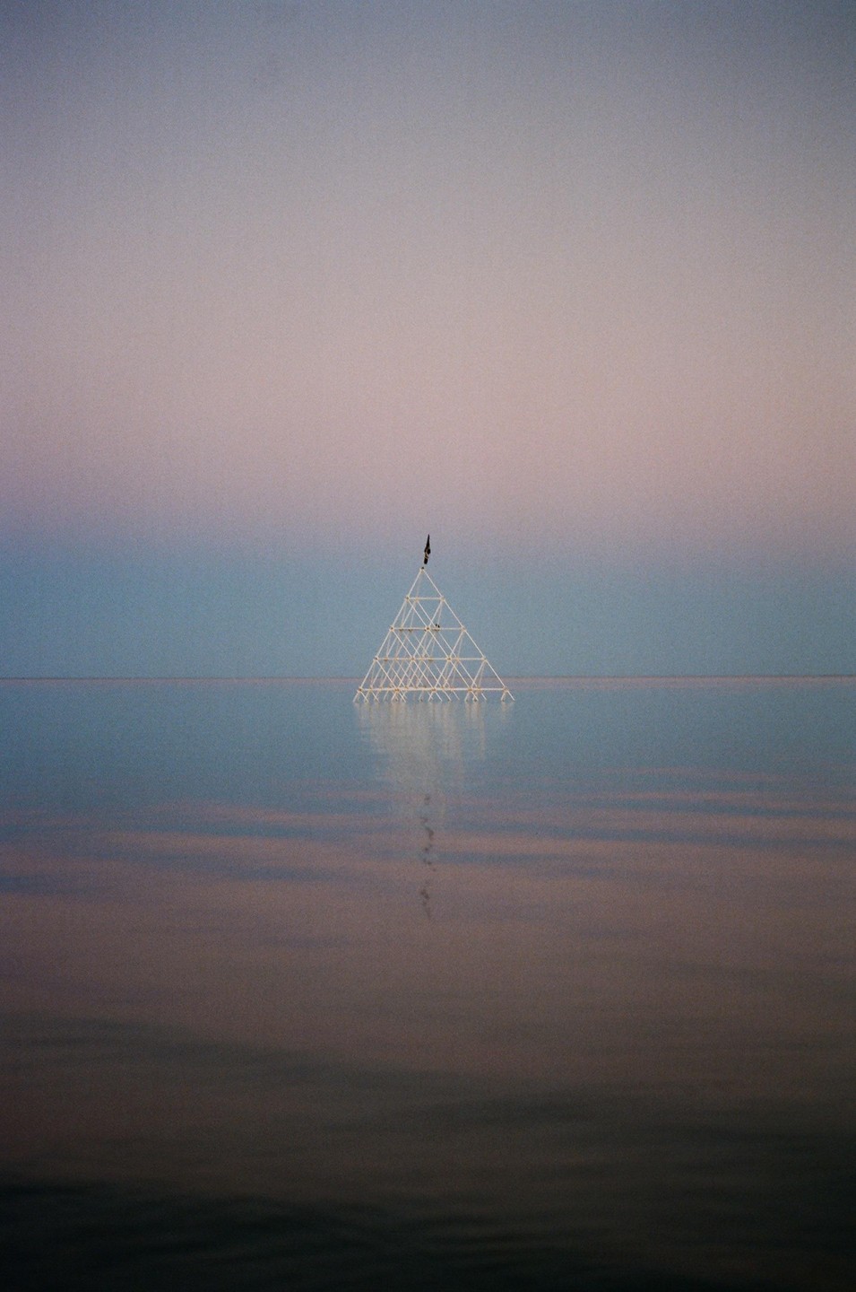 A triangular structure stands in the middle of a body of water under a gradient sky.