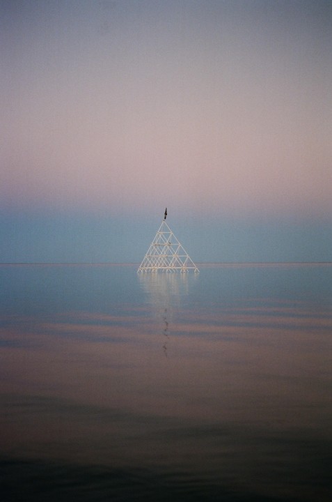 A triangular structure stands in the middle of a body of water under a gradient sky.