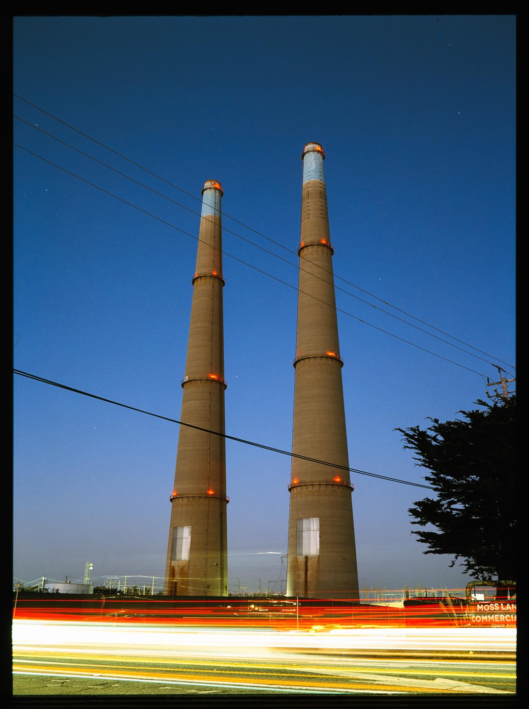 Two tall industrial chimneys stand against a deep blue night sky, with streaks of light from passing vehicles in the foreground.
