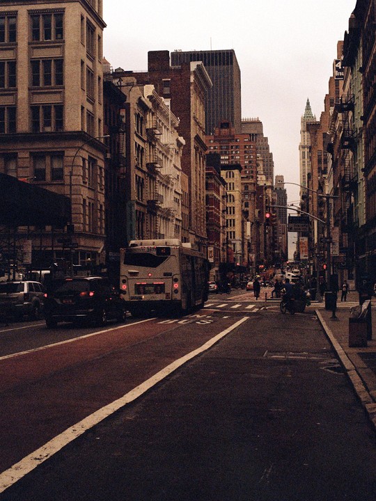 A bus and cars are stopped on a city street lined with tall buildings under an overcast sky.