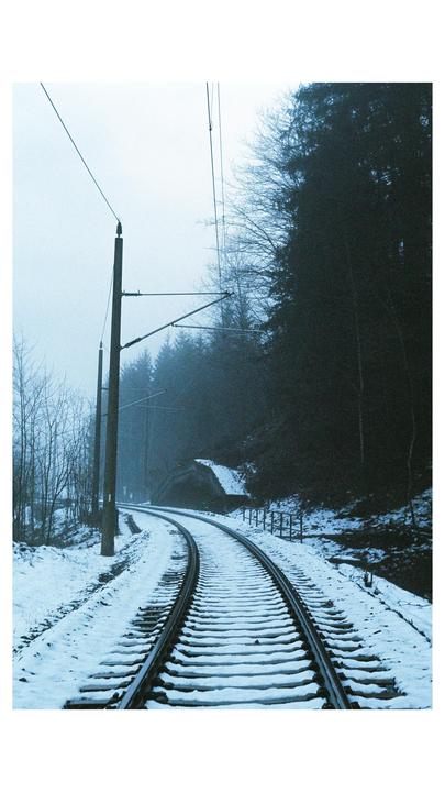 A snow-covered railway track curves through a forest with overhead power lines.
