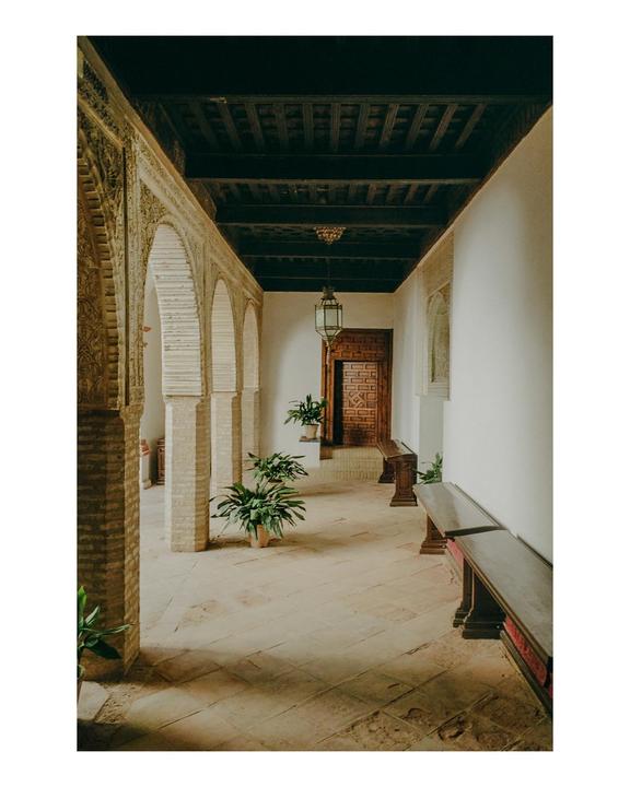 A long hallway with arches, benches, and potted plants leads to a carved wooden door.