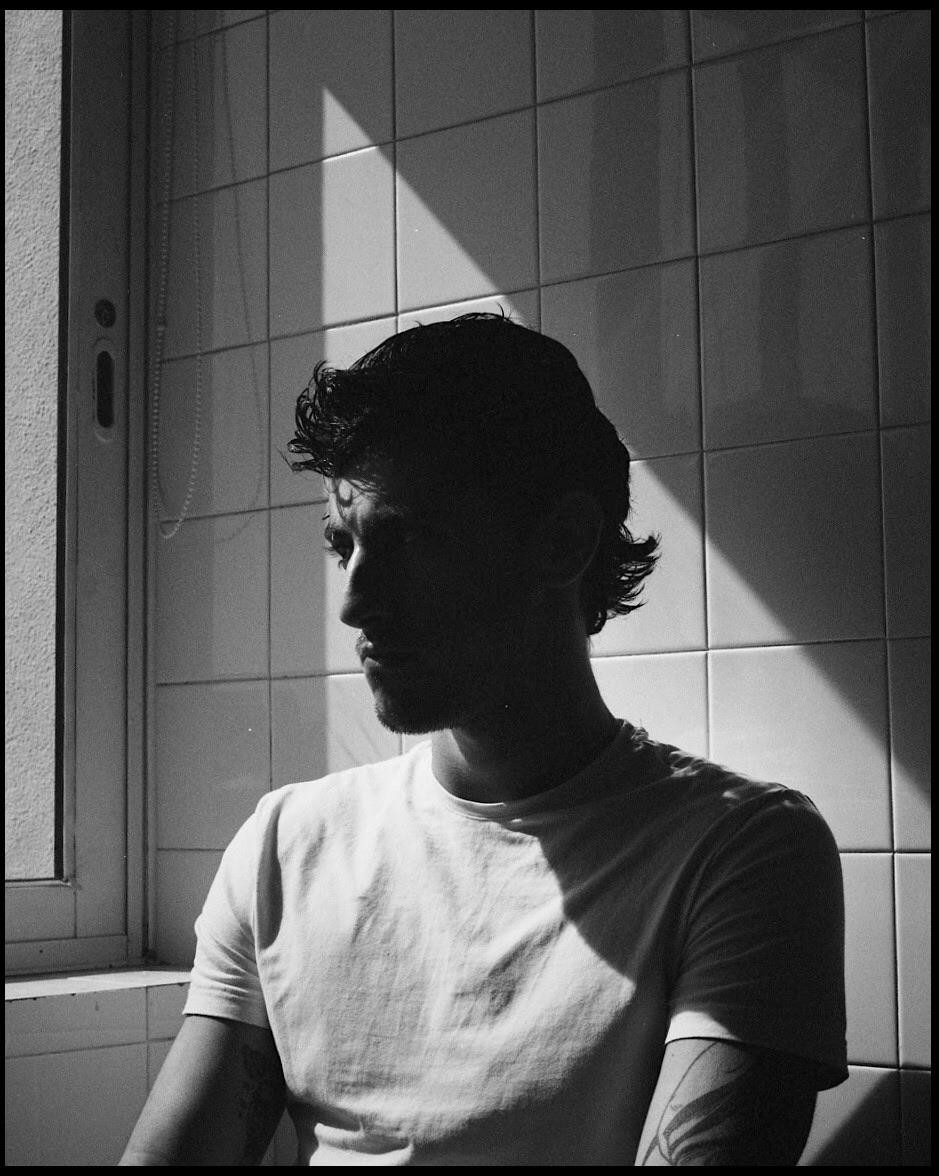 A man in a white t-shirt sits in a room with tiled walls, illuminated by a strong diagonal shadow.