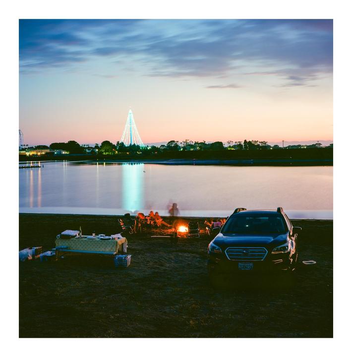 A group of people sit around a bonfire on a beach next to a lake with a large illuminated structure in the distance at dusk.