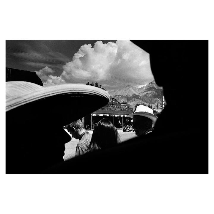 A group of people are visible in silhouette with mountains and a building in the background under a cloudy sky.