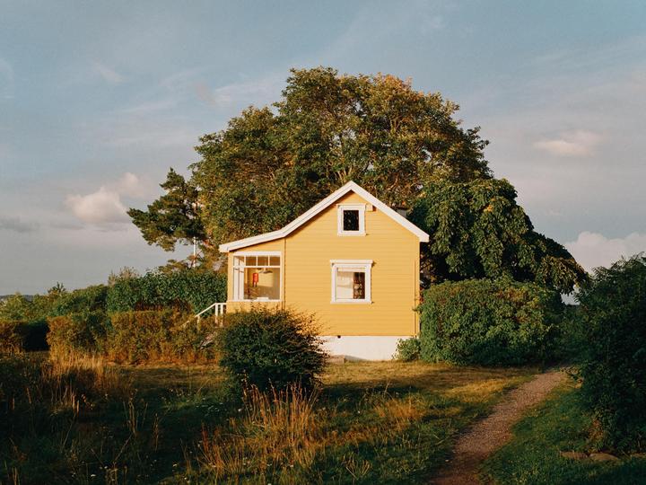 A yellow house with white trim is nestled among green trees and bushes under a partly cloudy sky.