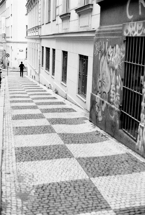 A man walks up a checkered cobblestone street lined with buildings, one of which has graffiti.