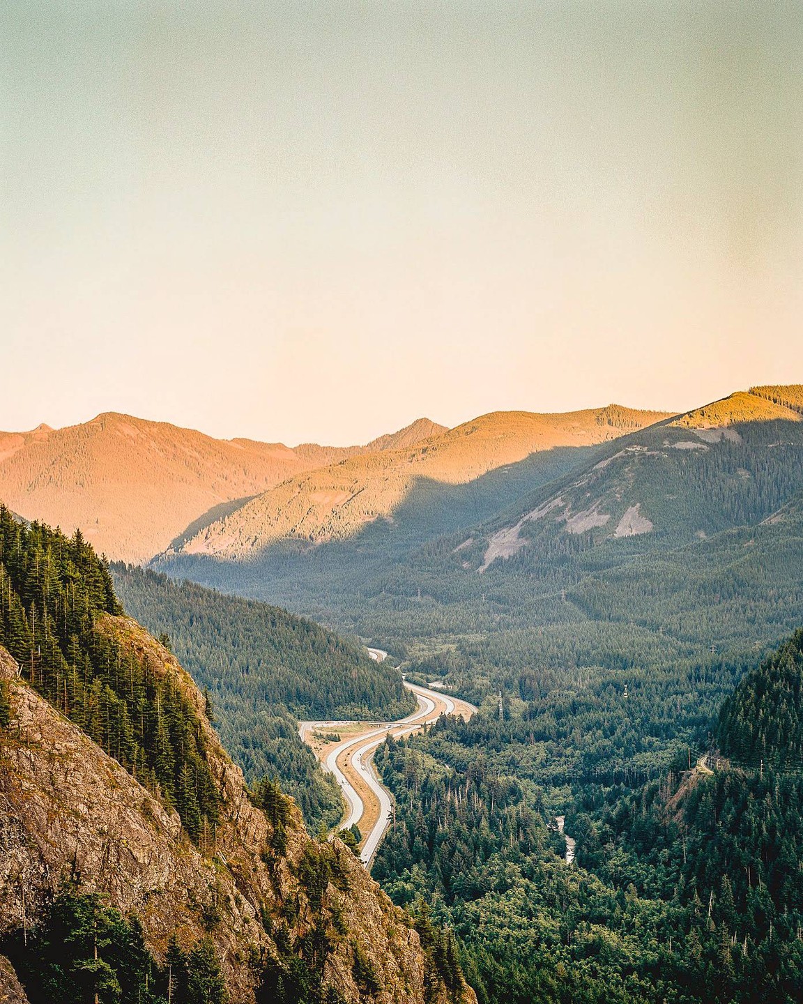 A winding road cuts through a valley surrounded by mountains and dense forest.