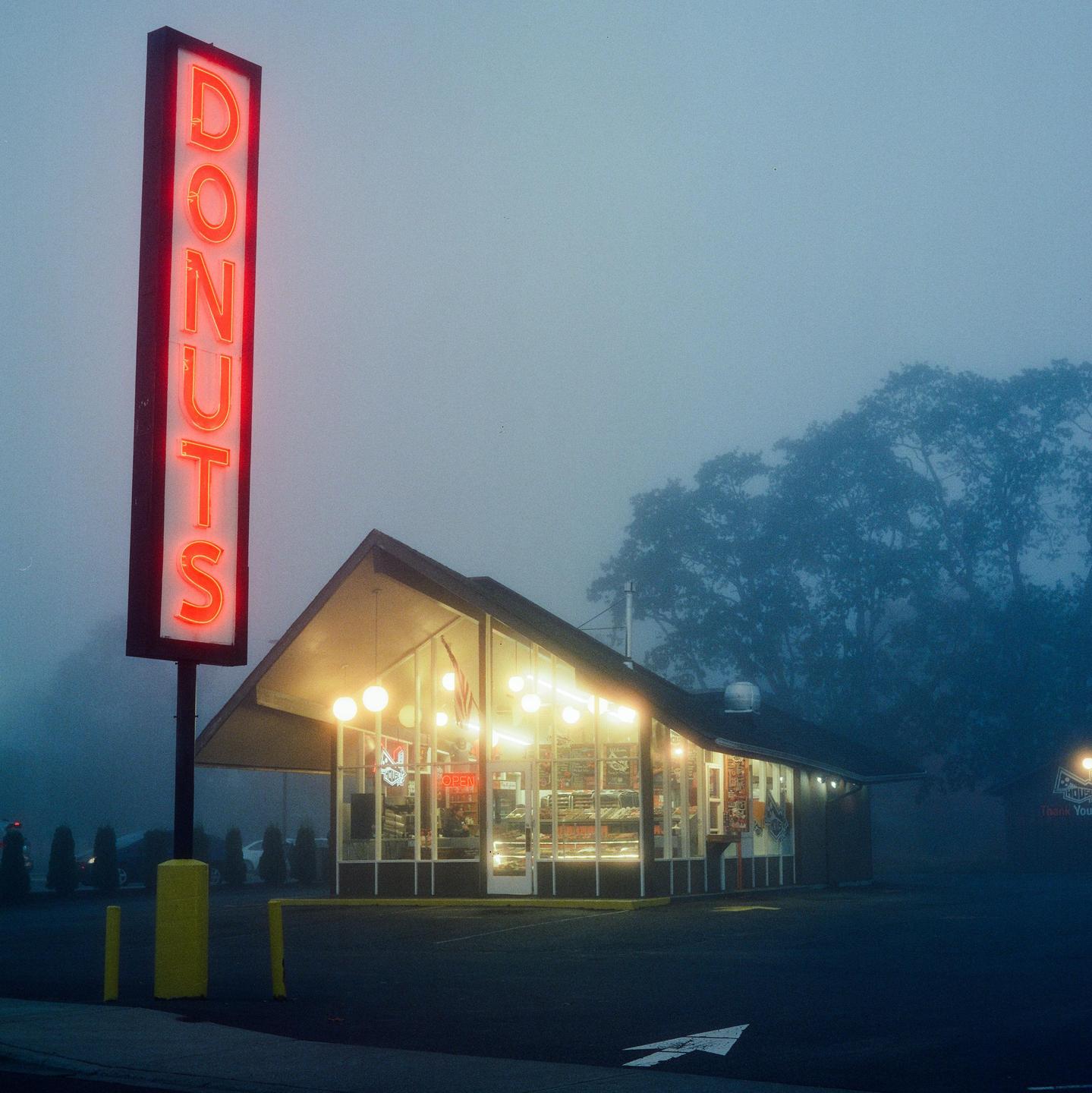 A neon sign for donuts stands next to a brightly lit donut shop on a foggy night.