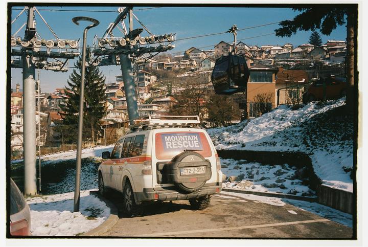 A mountain rescue vehicle is parked on a snowy road next to a ski lift with a town and mountains in the background.