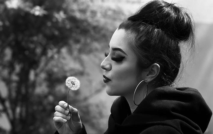 A woman with a bun in her hair holds a dandelion to her mouth.