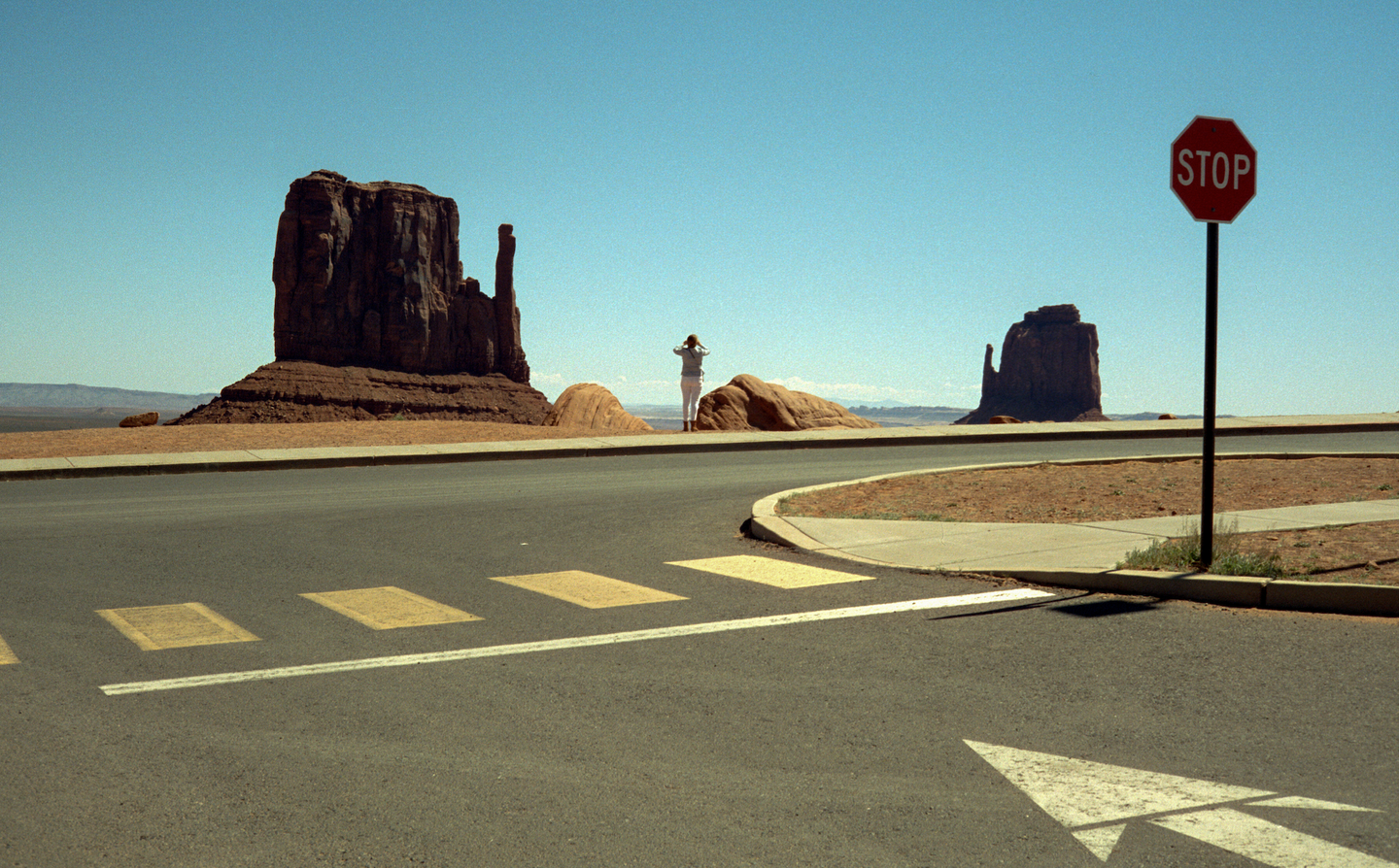 A woman stands on a dirt path looking at large rock formations in a desert landscape with a stop sign and road in the foreground.