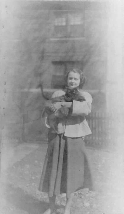 A woman holds a black cat in her arms while standing outside a building with a fence.