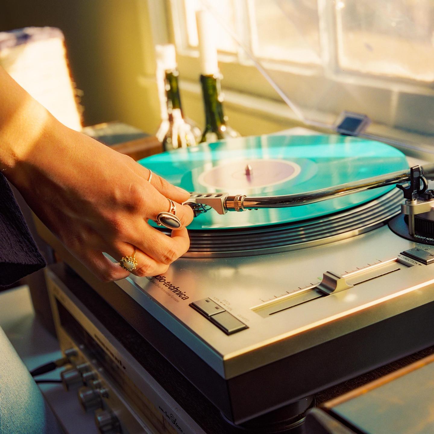 A hand with rings places a needle on a turquoise record on a turntable.