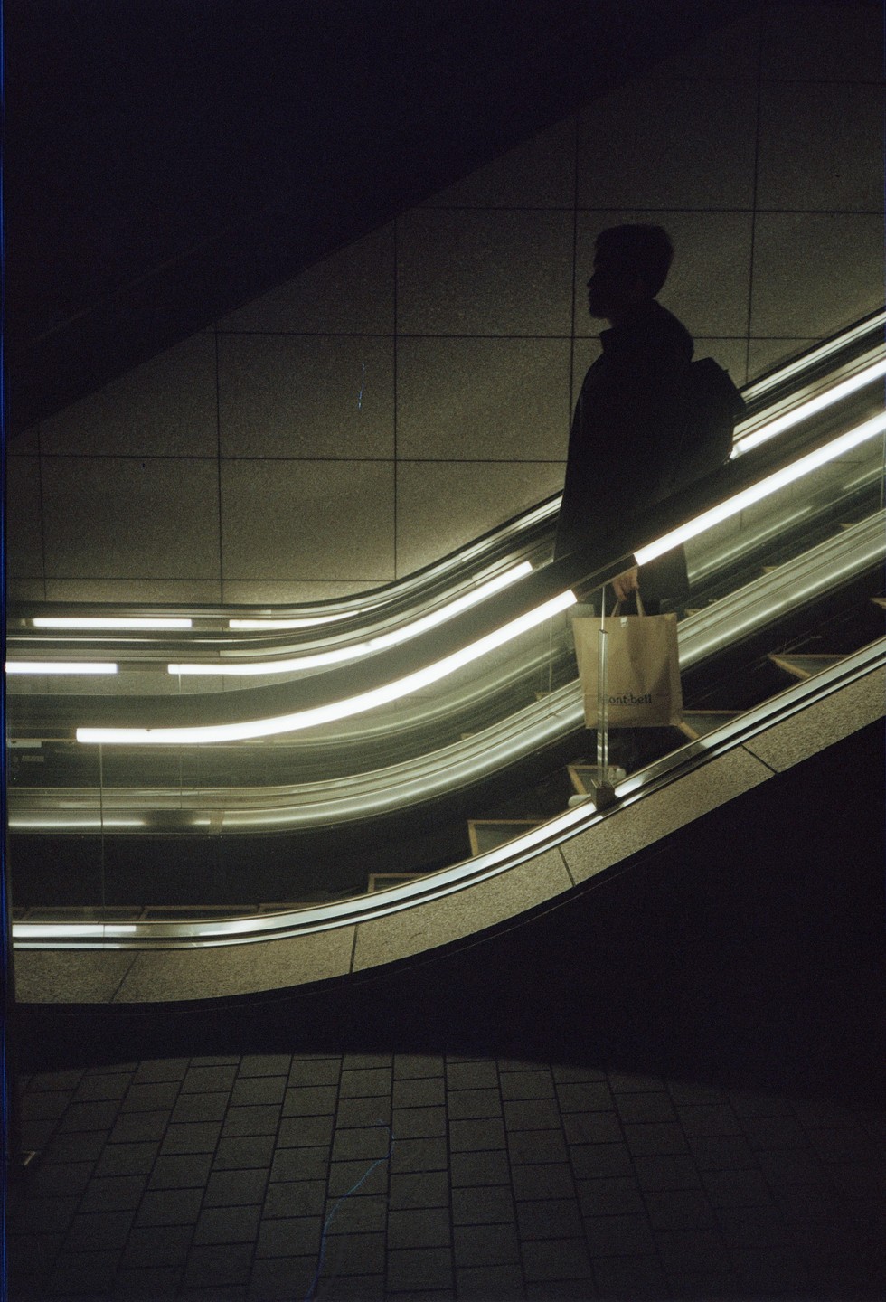 A person with a backpack and a shopping bag stands on an escalator with bright lights.