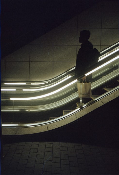 A person with a backpack and a shopping bag stands on an escalator with bright lights.