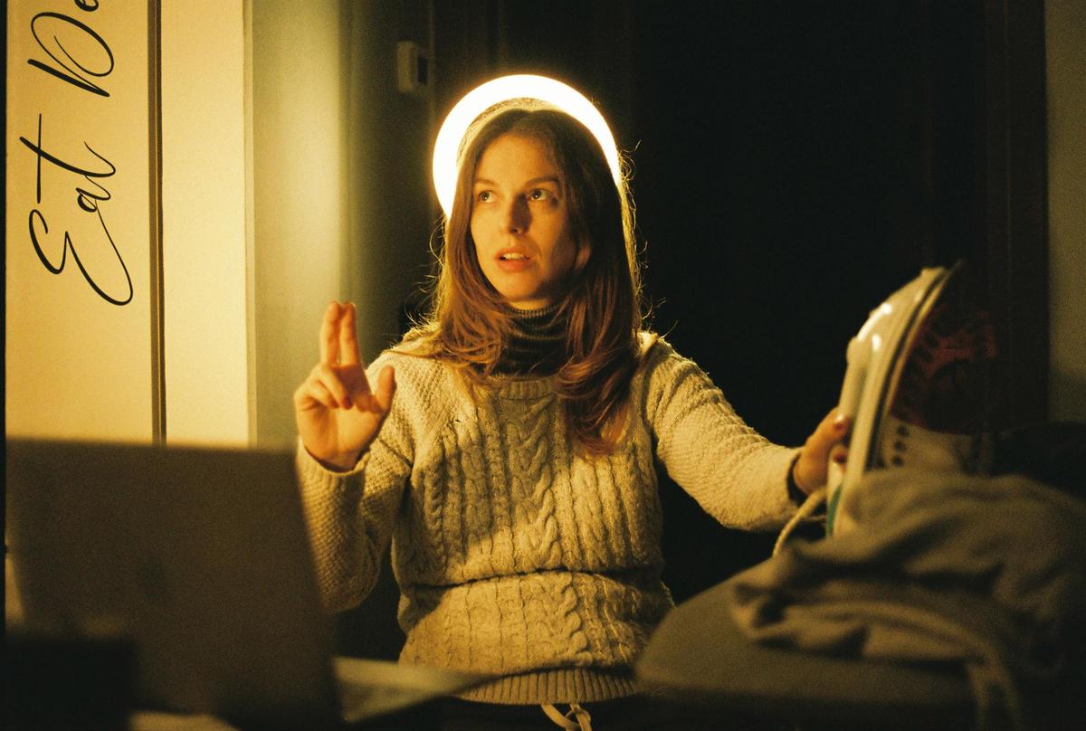 A woman with long hair and a cable-knit sweater holds an iron and gestures with her other hand, with a ring light behind her head.