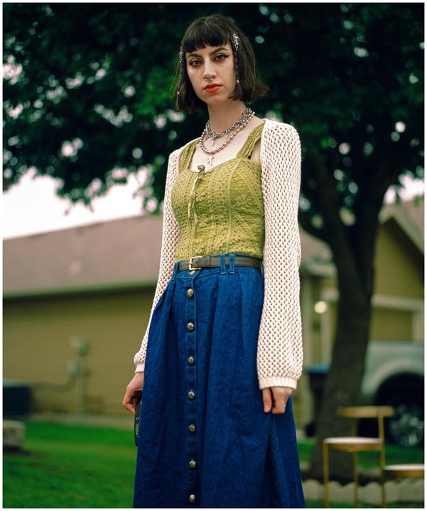 A woman with dark hair and bangs stands outdoors, wearing a green top, a blue skirt, and a white crocheted cardigan.