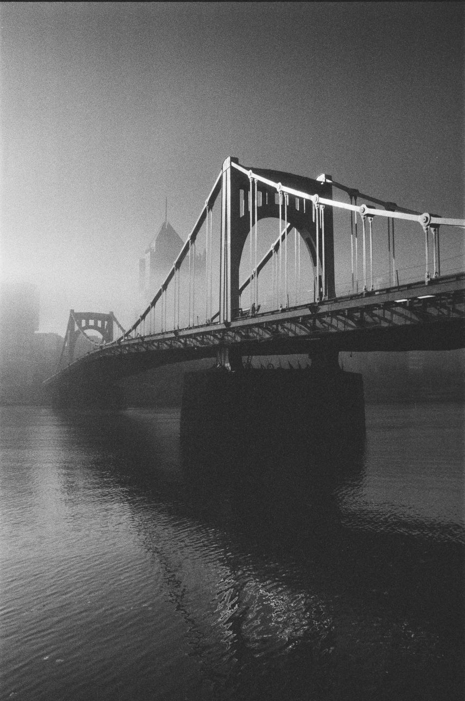 A suspension bridge spans a body of water with a hazy city skyline in the background.