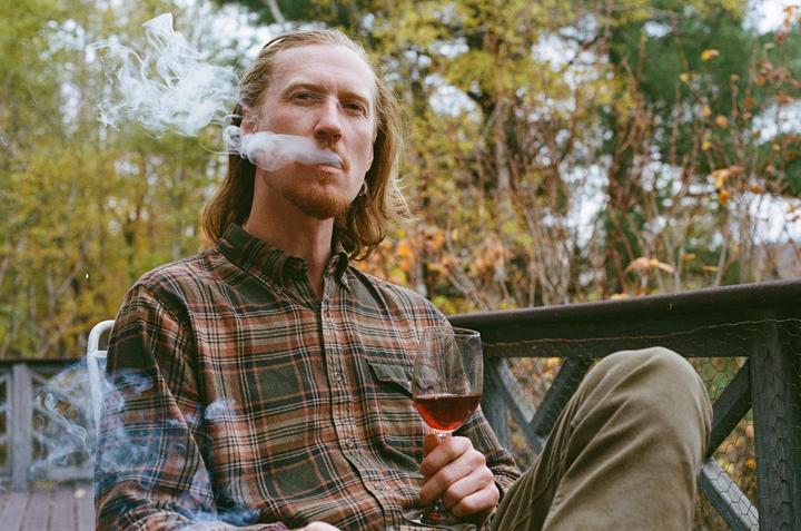A man with long hair and a beard sits on a porch, holding a glass of red wine and exhaling smoke.