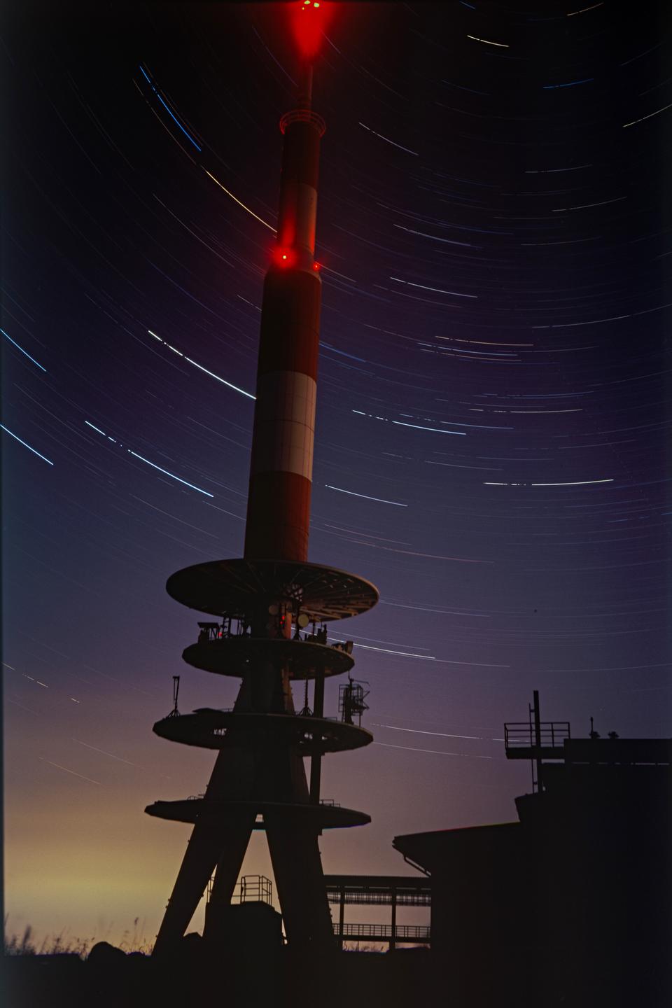 A red and white tower stands against a night sky filled with star trails.
