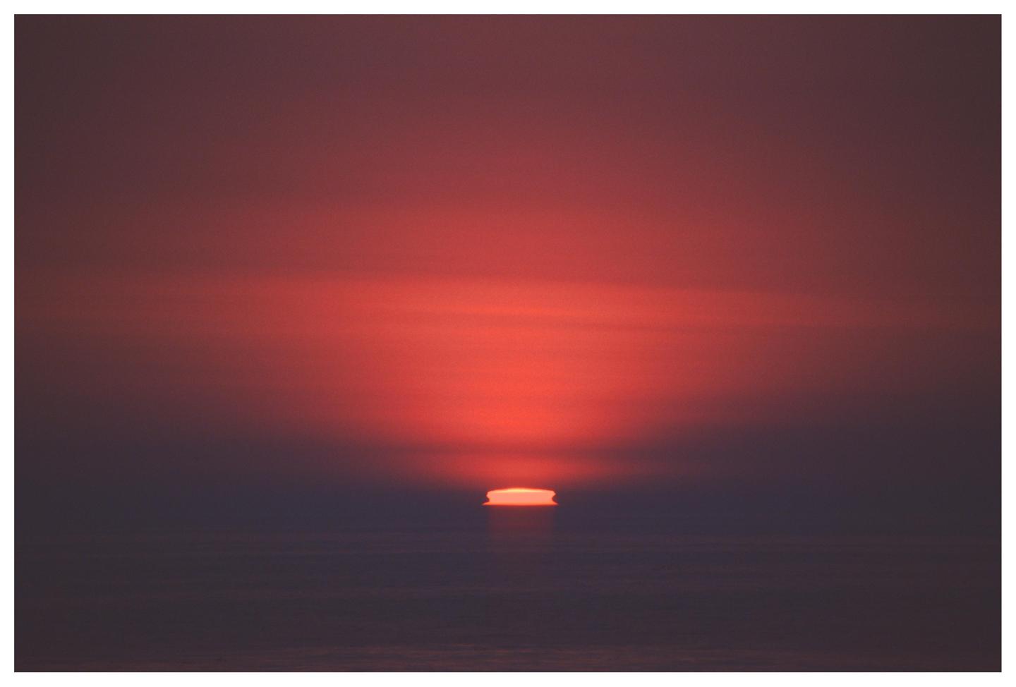 The sun dips below the horizon, casting a red glow across the sky and sea.