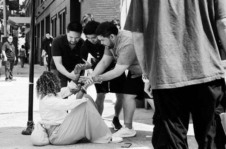 A woman sits on the sidewalk surrounded by several men who are helping her.