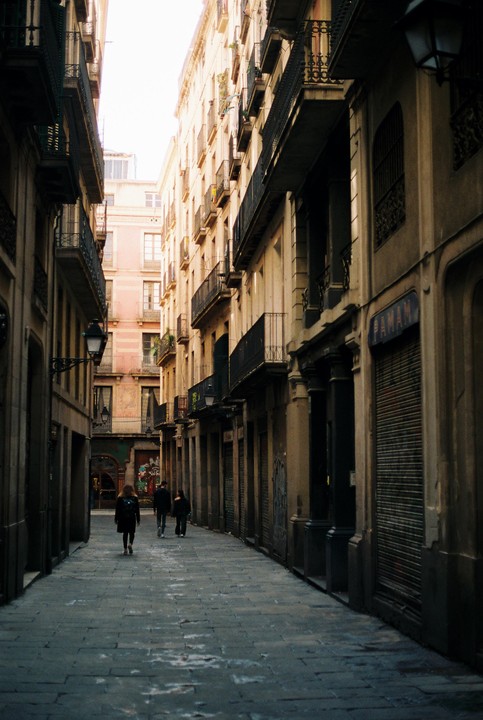 Three people walk down a narrow alley lined with tall buildings and closed storefronts.