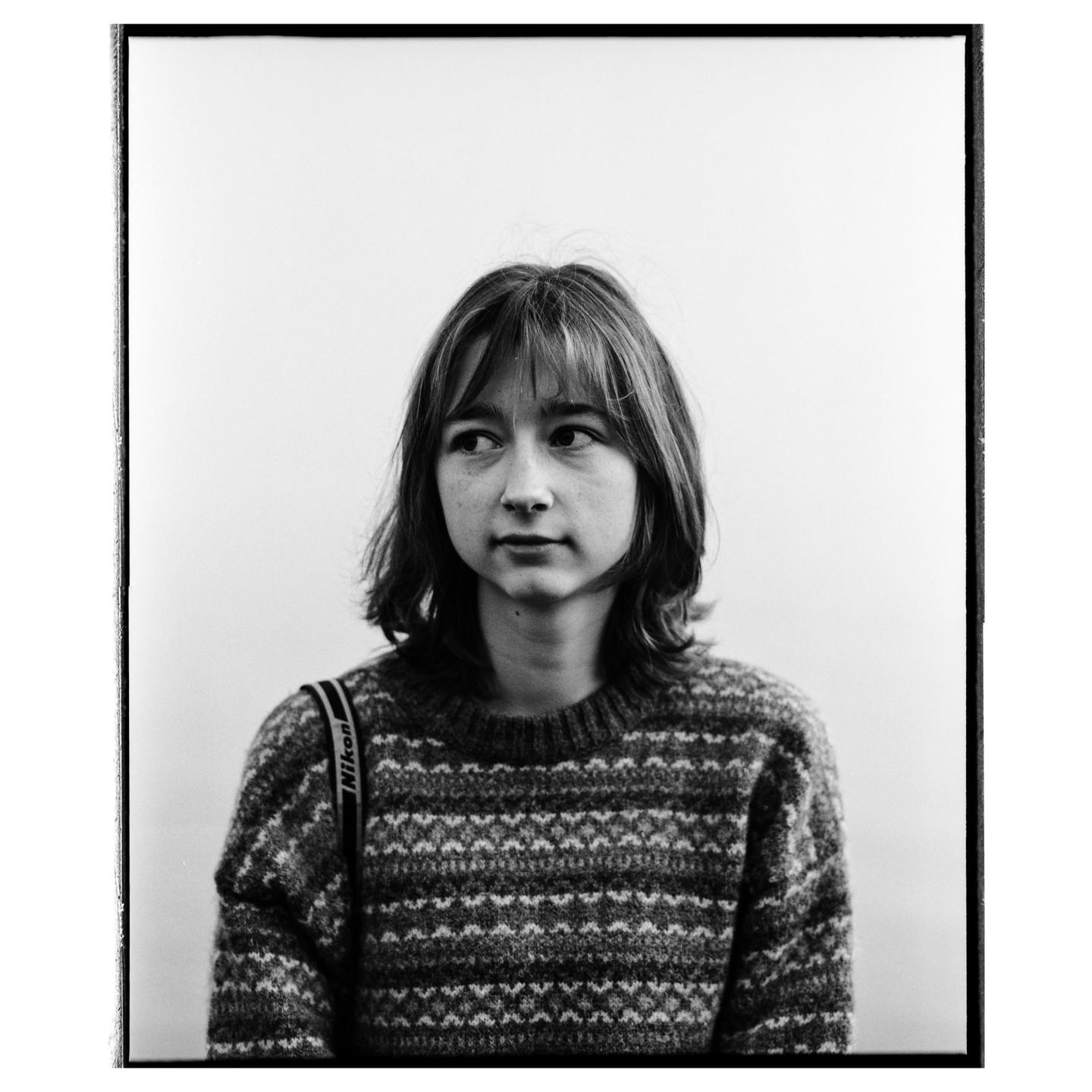 A young woman with bangs and a patterned sweater looks to the side.