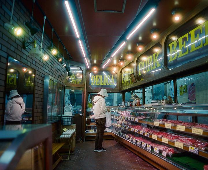 A person wearing a bucket hat stands in a brightly lit butcher shop, looking at cuts of meat in a refrigerated display case.