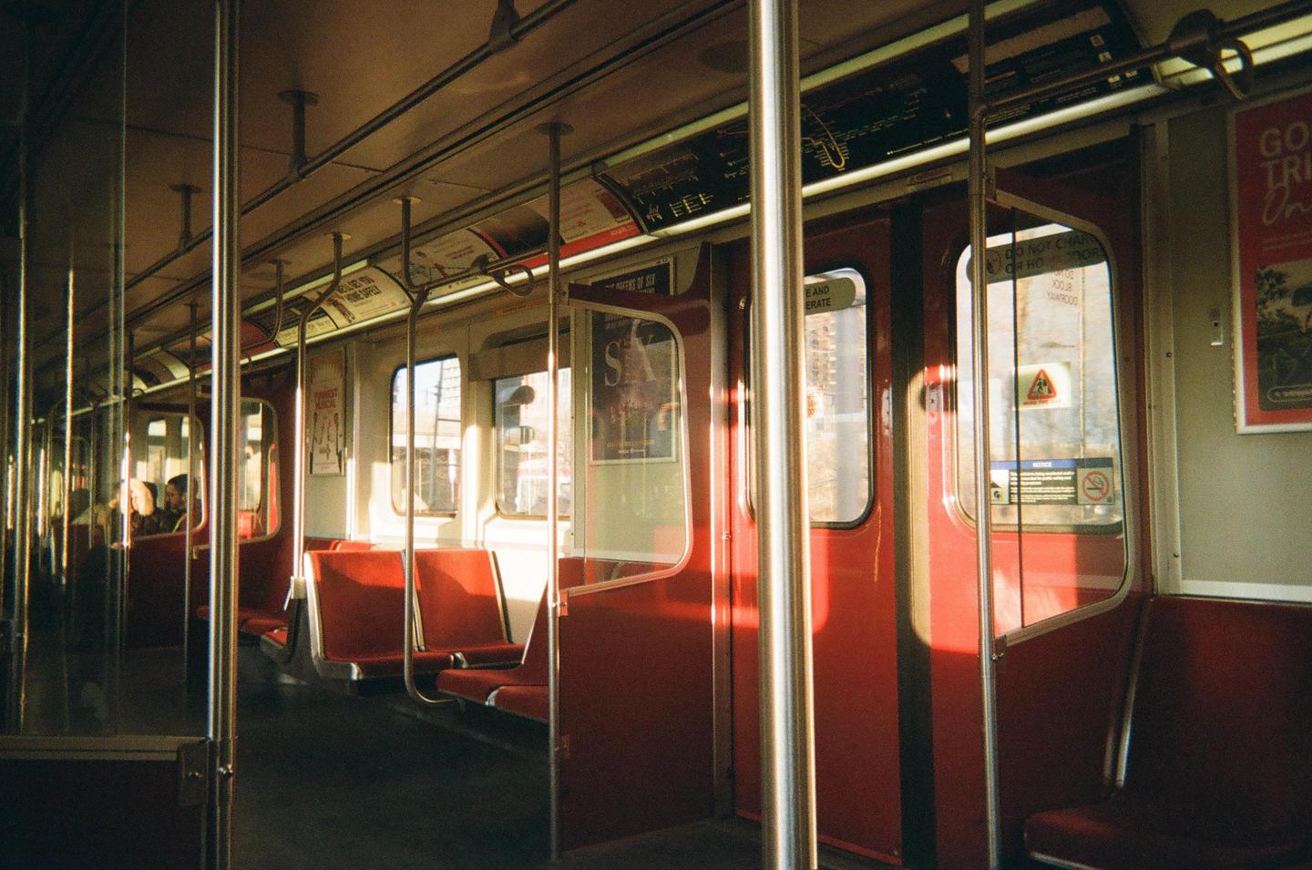 Sunlight streams through the windows of a subway car with red seats and a few passengers.