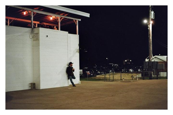 A man in a cowboy hat leans against a white building at night, with a parking lot and utility pole visible in the background.