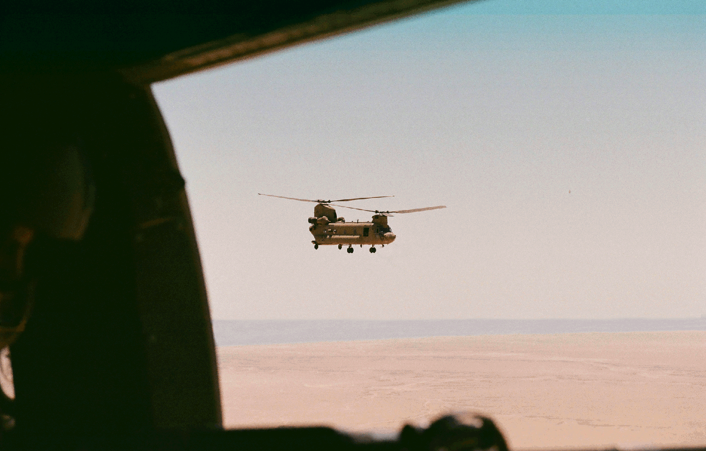 A helicopter flies over a desert landscape, viewed from inside another aircraft.