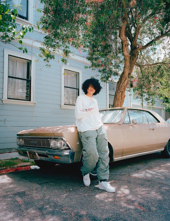 A person with curly hair leans against a gold vintage car parked on a street in front of a light blue house.