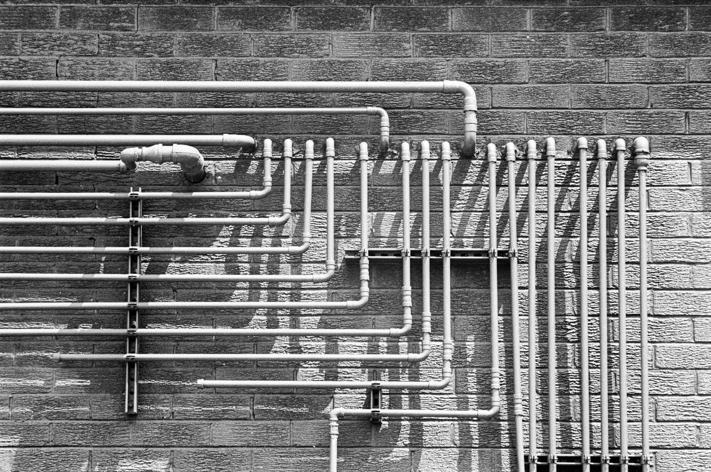 A network of pipes runs horizontally and vertically across a brick wall, casting shadows.